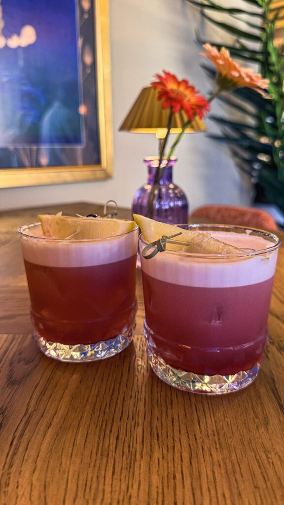 2 pink drinks with creamy top, garnished with lemon on a table with fresh flowers behind