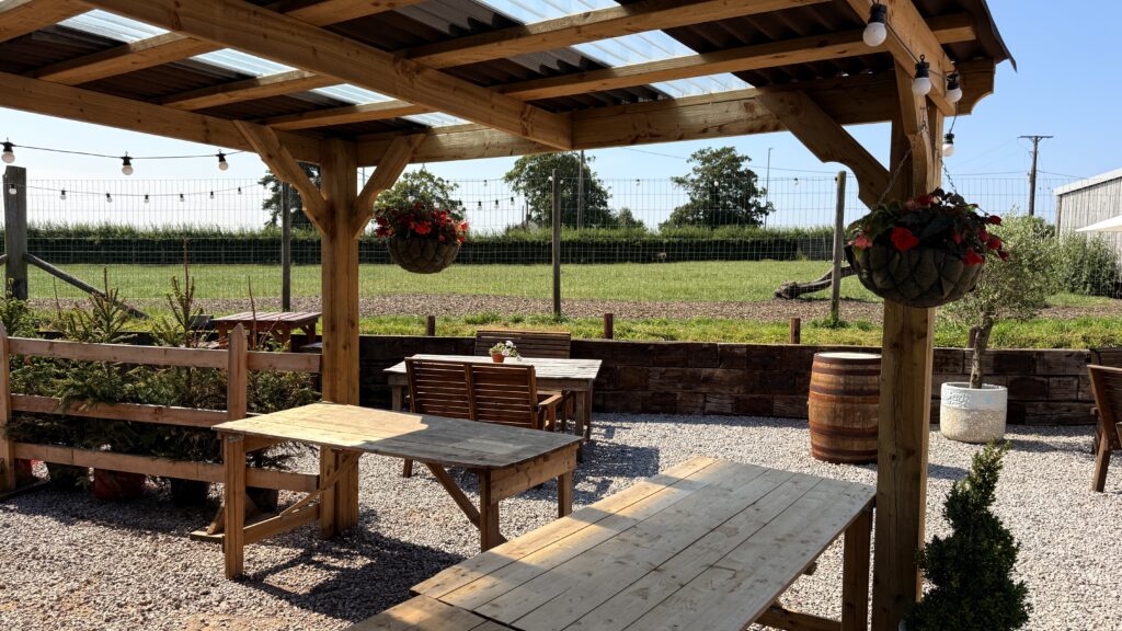 Wooden pergola seating area at Oakleys Farm, with string lights, hanging flower baskets and a barrel table.